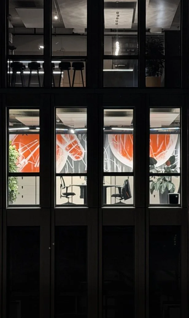Interior office space seen through large glass windows, with two chairs and a table, decorated with large abstract orange and white artwork and some greenery.