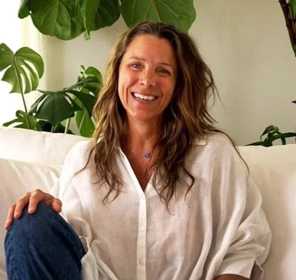 A woman with wavy, light brown hair smiling outdoors with green foliage in the background.