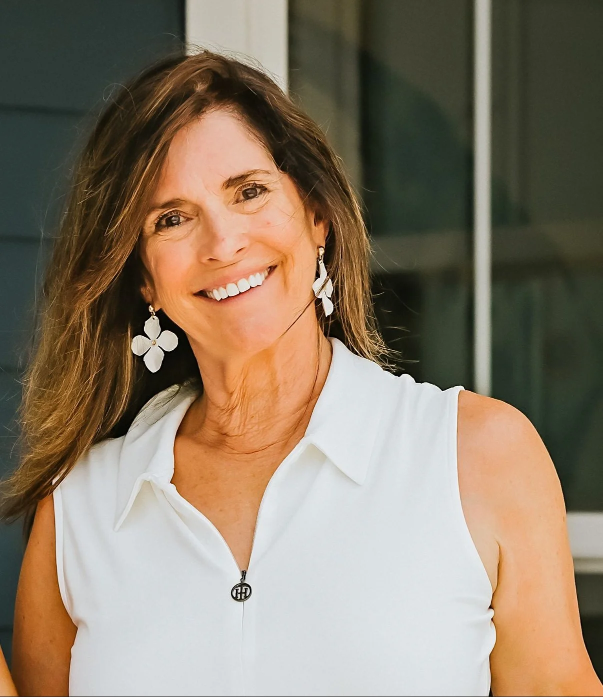 Smiling woman with long brown hair, wearing white sleeveless top, floral earrings, standing outdoors in front of a building.