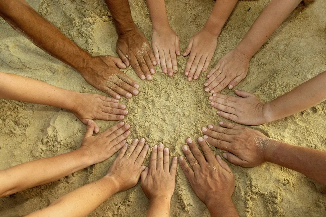 Multiple hands placed on the sand, forming a heart shape.