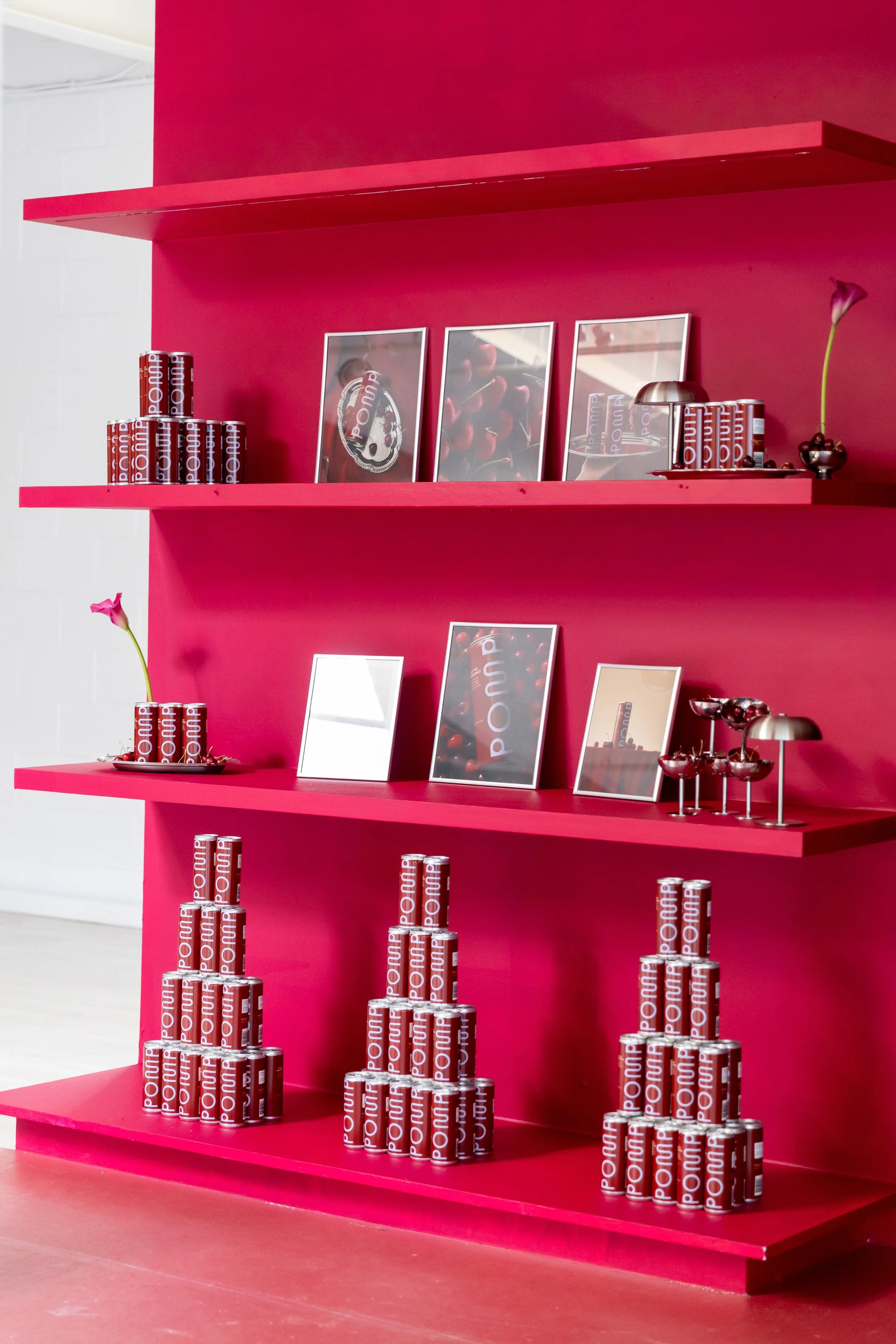 Display shelf in pink with cans labeled 'POUN'. Three framed pictures, one of cherries, one of a pie, and one with abstract design. A single flower in a metal vase and a tray of cherries are also on the shelf.