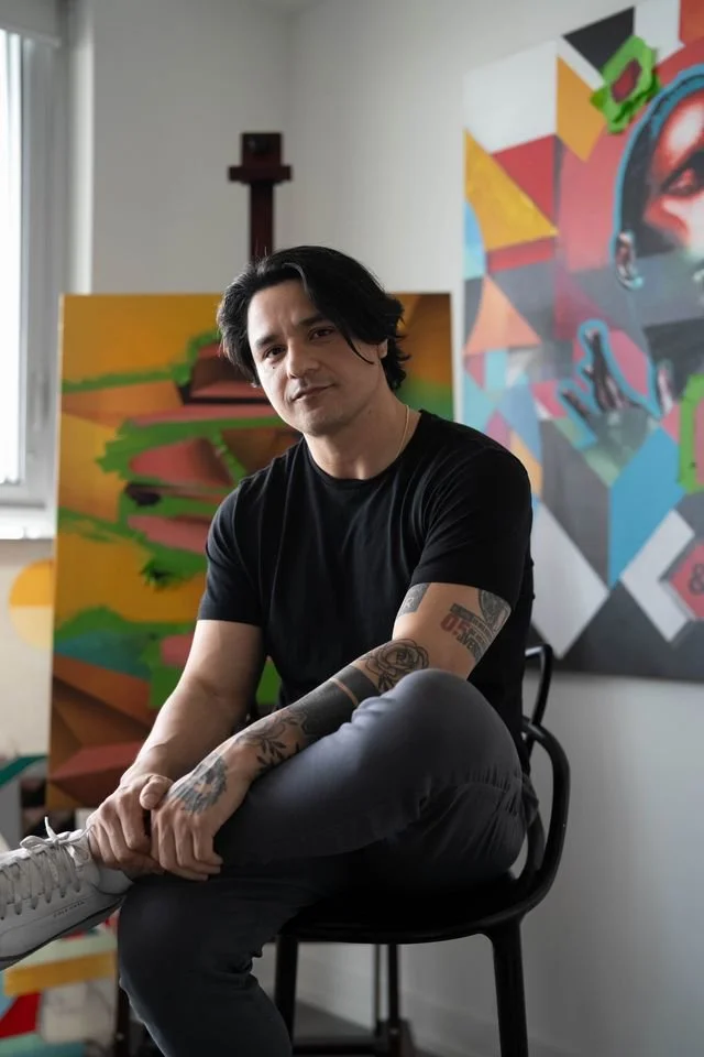 A man with black hair and tattoos on his arm, wearing a black t-shirt and gray jeans, sitting on a black chair in an art studio with colorful abstract paintings in the background.