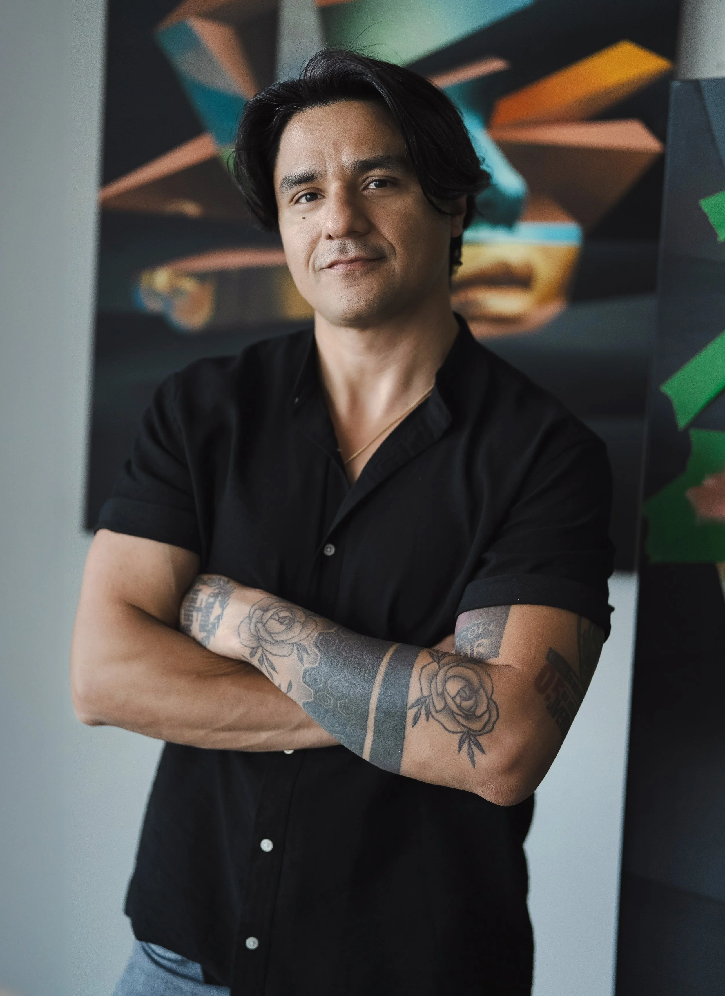 A man with black hair and tattoos on his arms standing with arms crossed in front of a colorful geometric background.