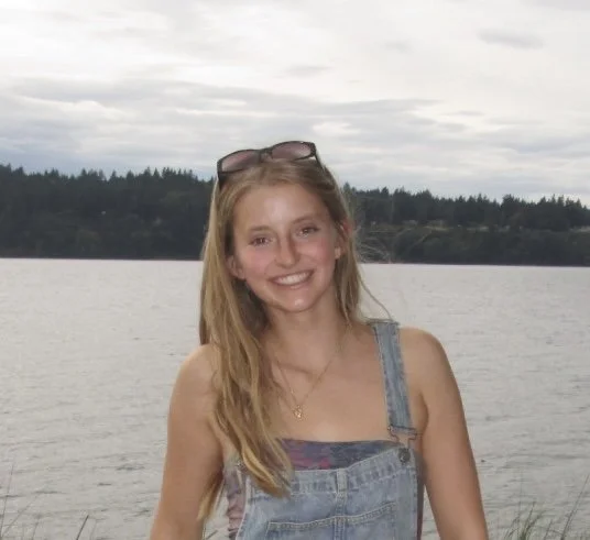 A young woman smiling outdoors near a body of water, with trees and cloudy sky in the background.