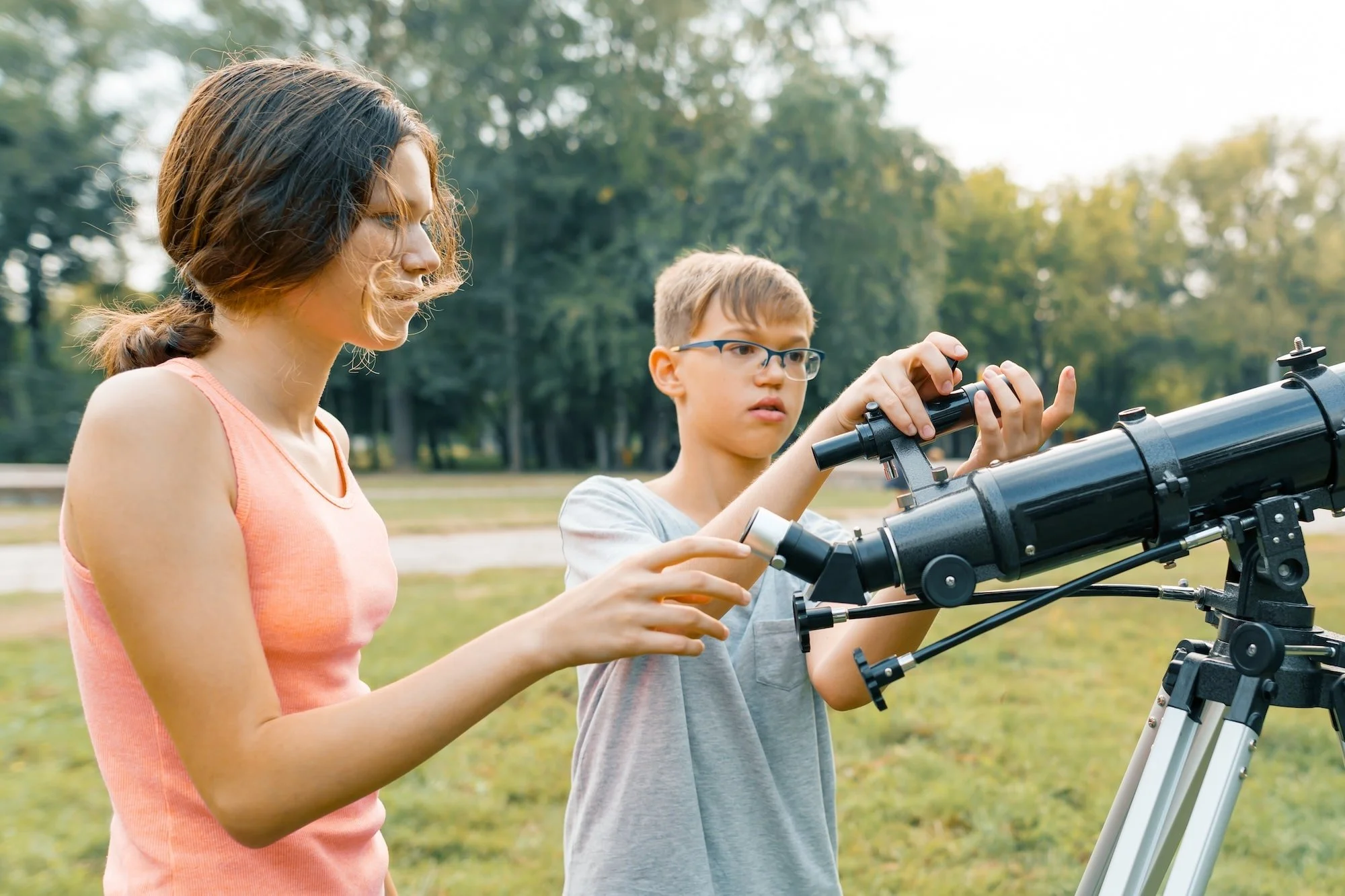 A young girl and boy outdoors looking through a telescope in a park.