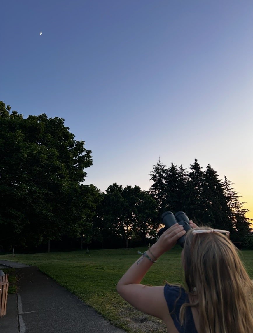A girl with long blonde hair looking through binoculars in a park during sunset, with green trees and a sky with a crescent moon.