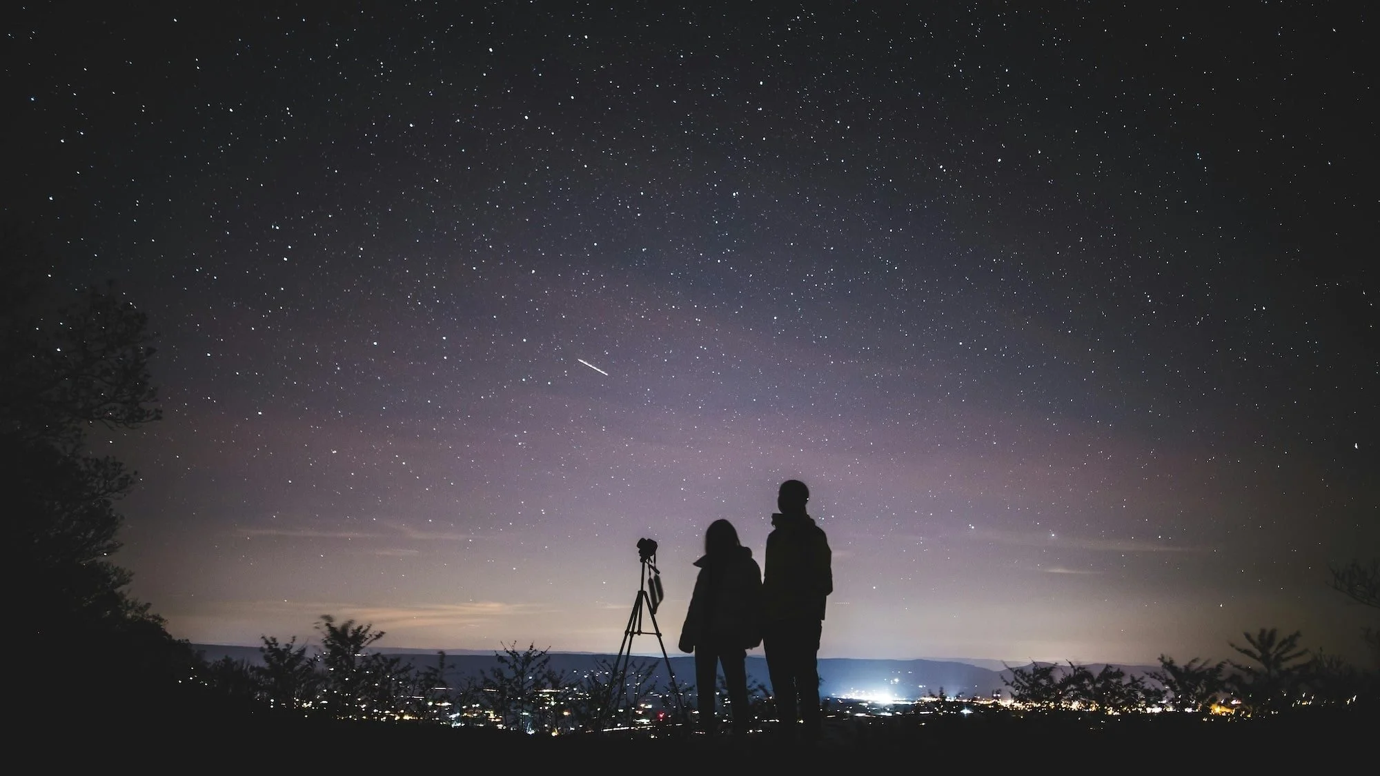 Silhouettes of two people observing the night sky with a telescope on a hilltop, stars and a streak of light across the starry sky, city lights below.