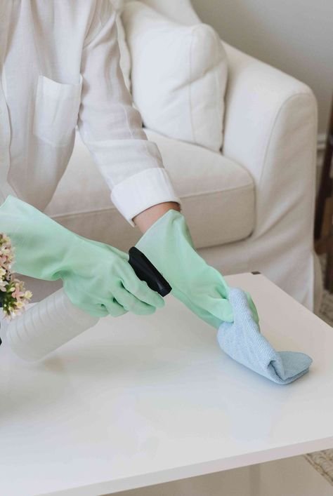 Person cleaning a white table with spray bottle and cloth, wearing green gloves.