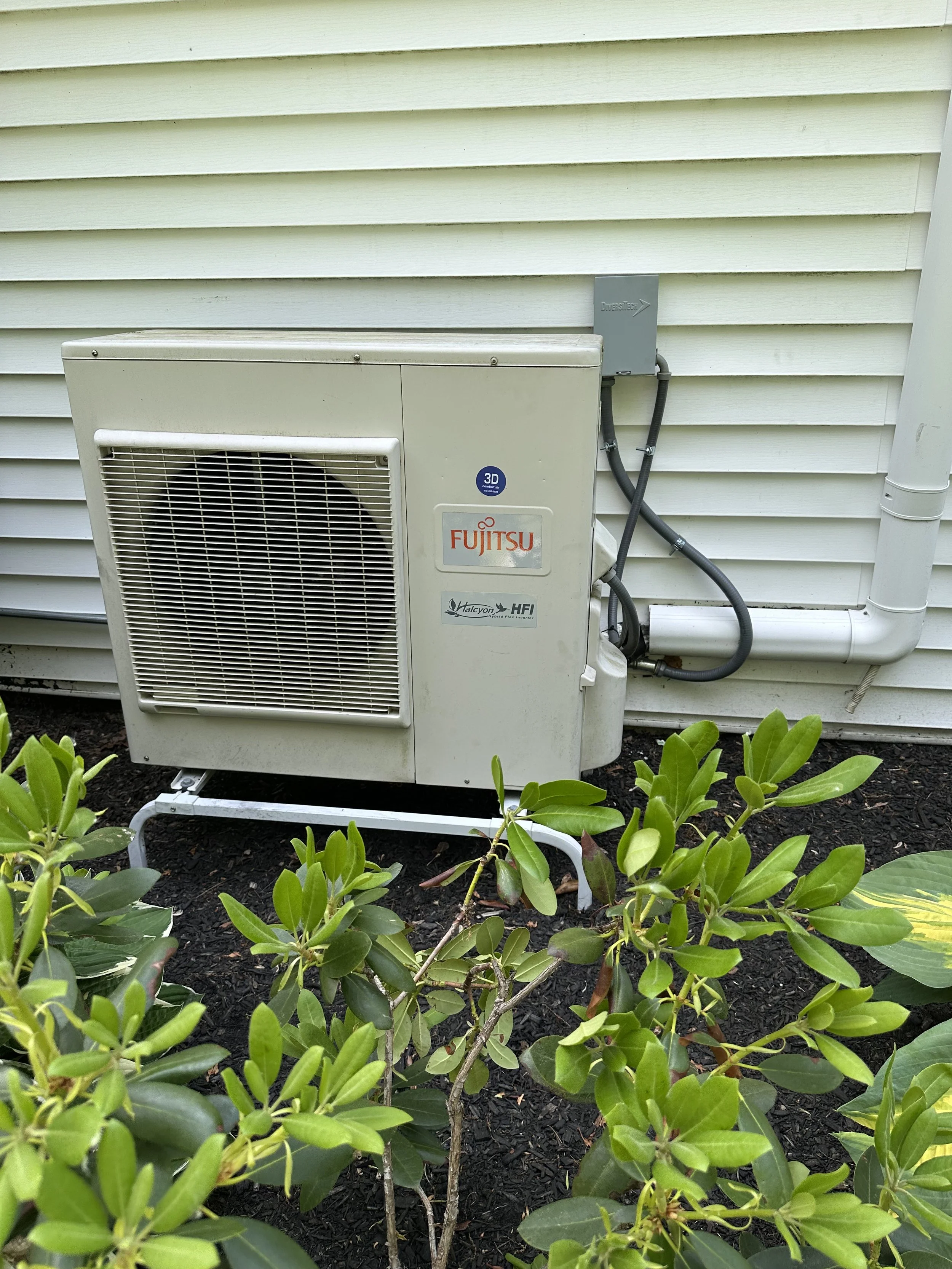 Outdoor air conditioning unit installed outside house with green shrubbery in front.