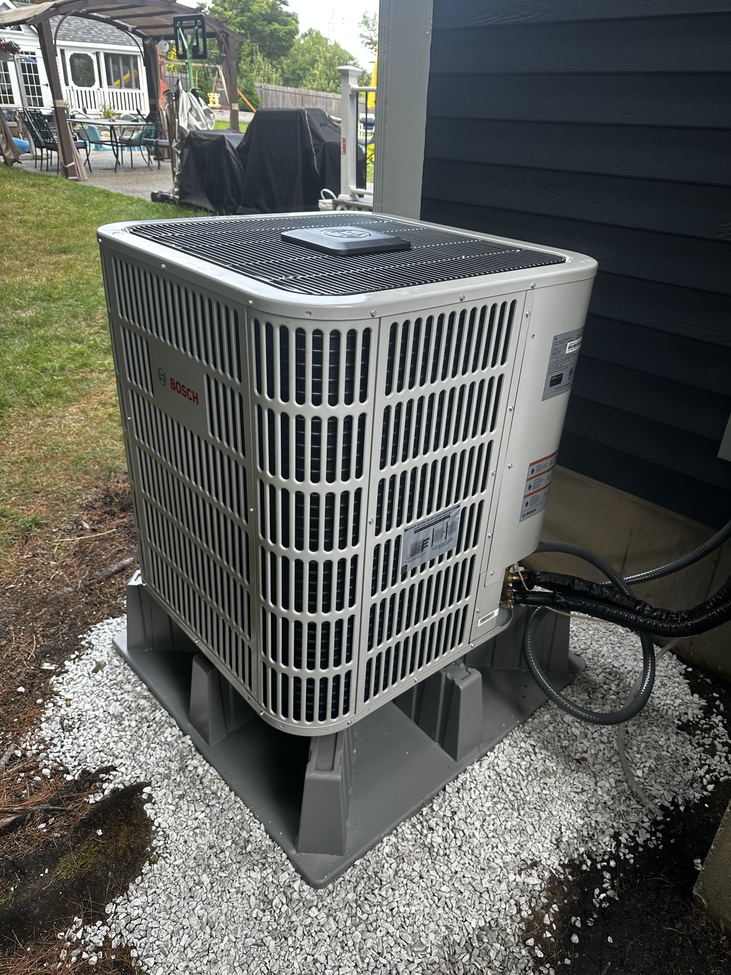 A Bosch air conditioning unit installed outside on a gray platform surrounded by gravel, next to a dark blue house wall.