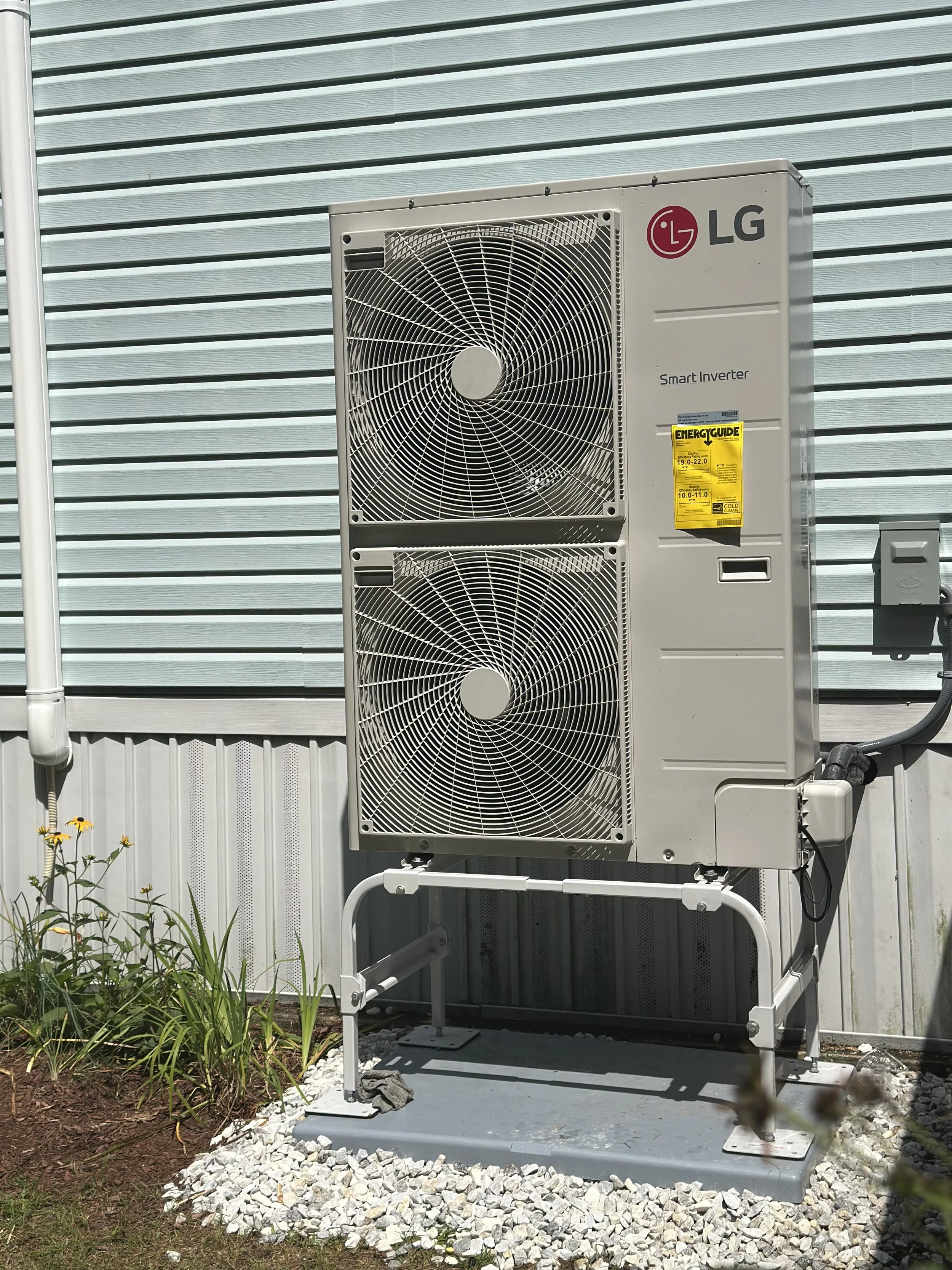 An outdoor air conditioning unit with LG branding, mounted on a metal stand with white gravel underneath, positioned beside a building with metal siding.