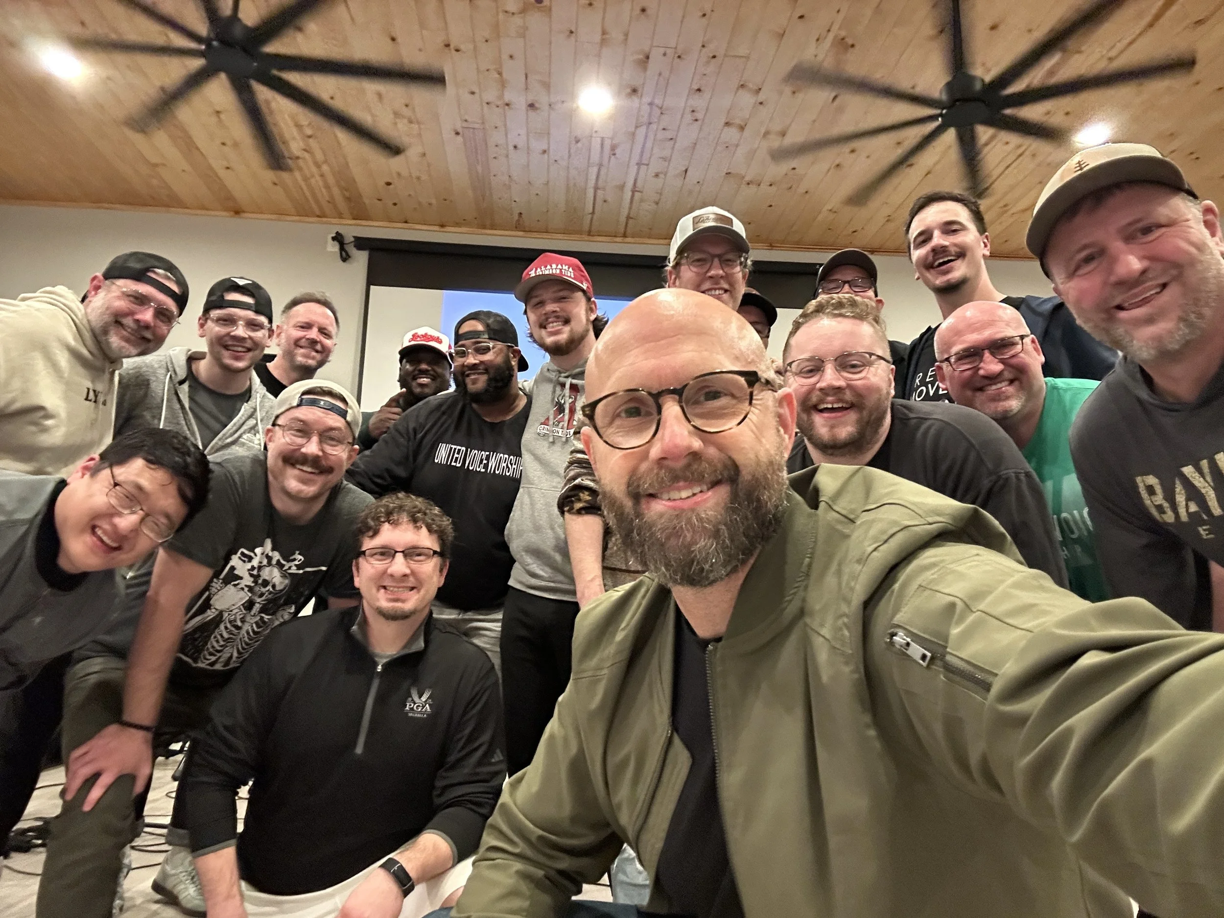 Group of smiling men taking a selfie in a room with a wooden ceiling, some wearing caps and glasses.