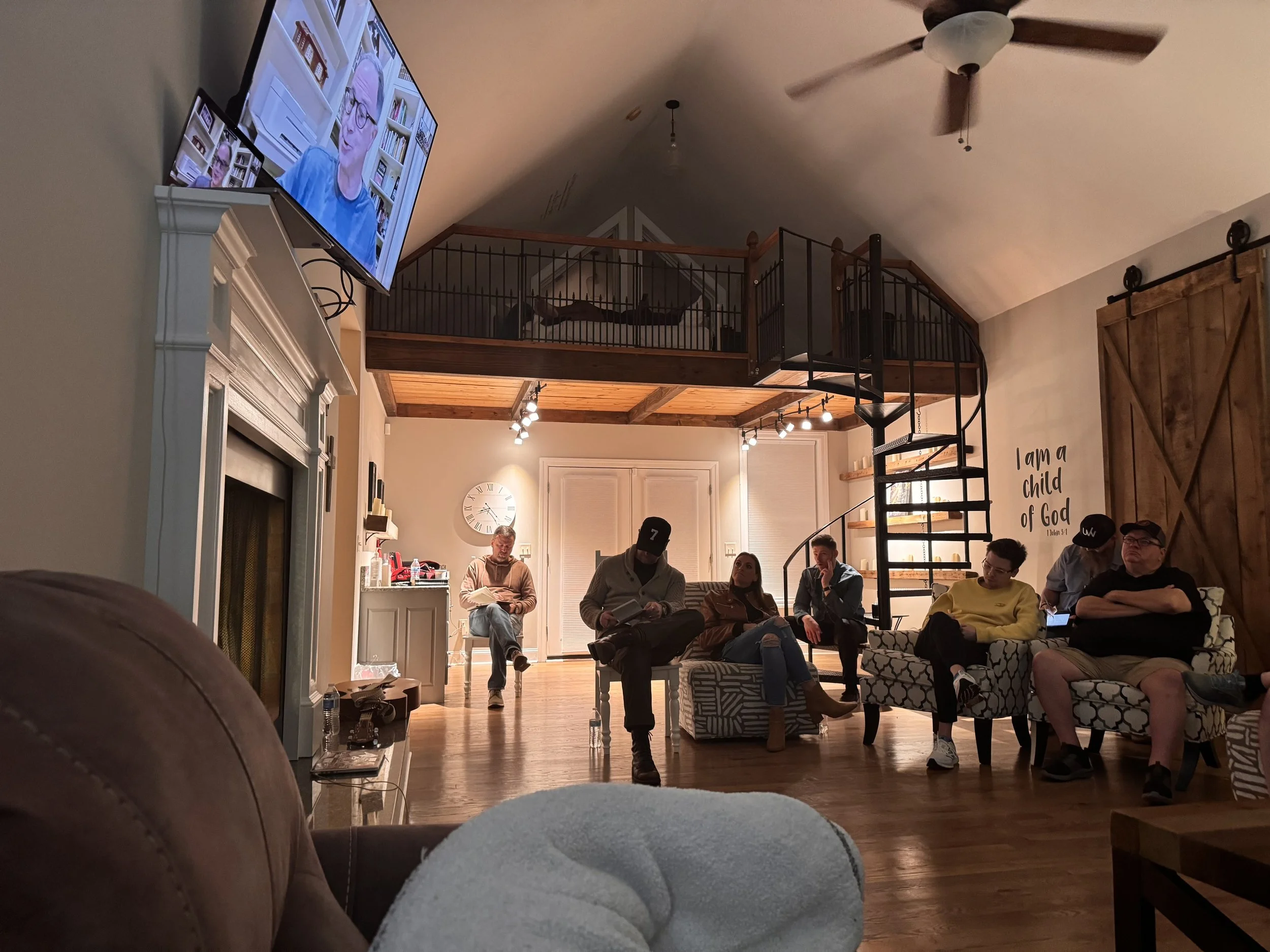 Living room with a group of people sitting on couches and chairs, a large wall clock, a television showing a woman speaking, and a loft area with wooden railing and a ceiling fan.