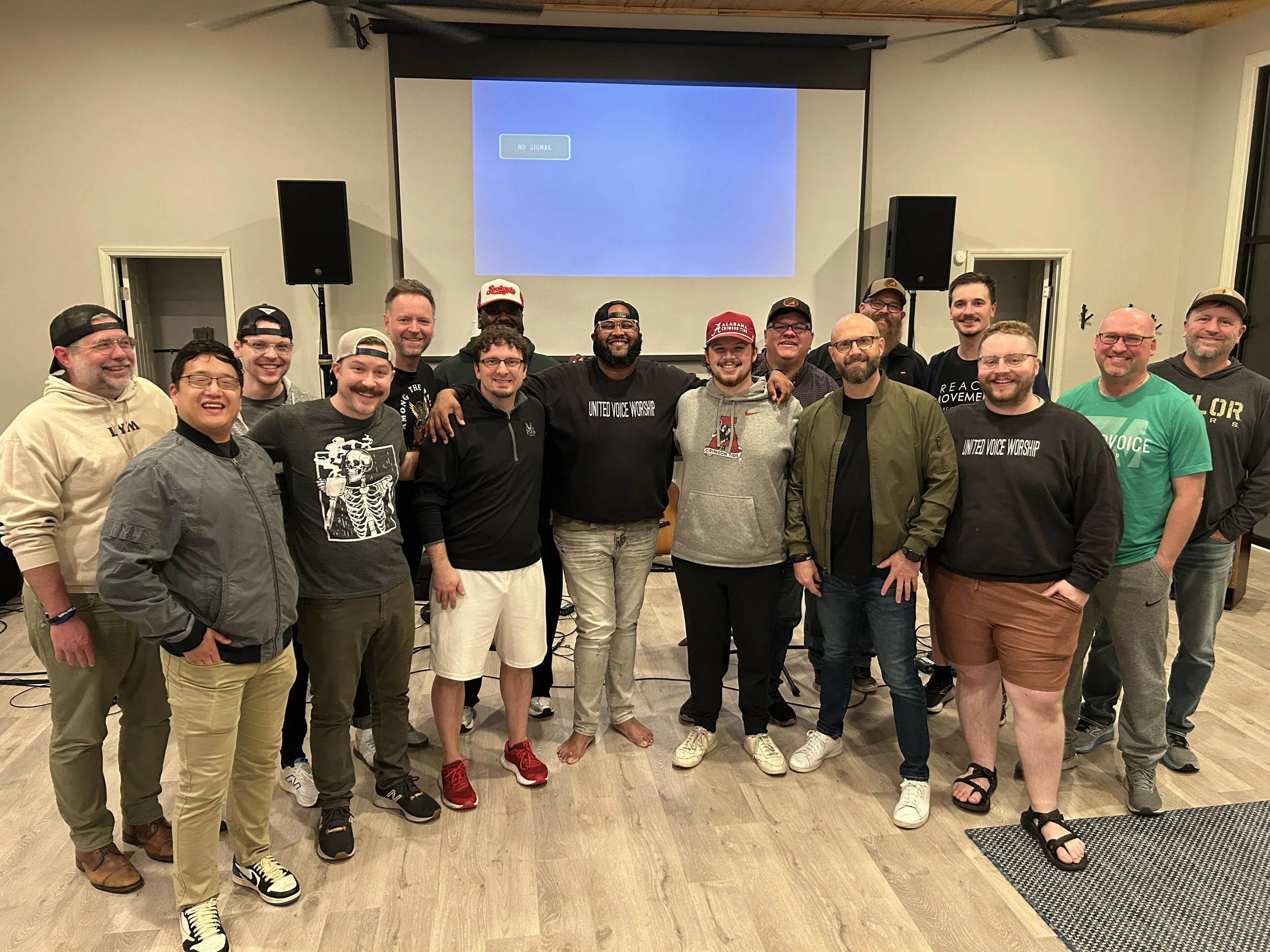 Group of 16 smiling men standing in a room with a blank TV screen behind them, some with arms around each other.