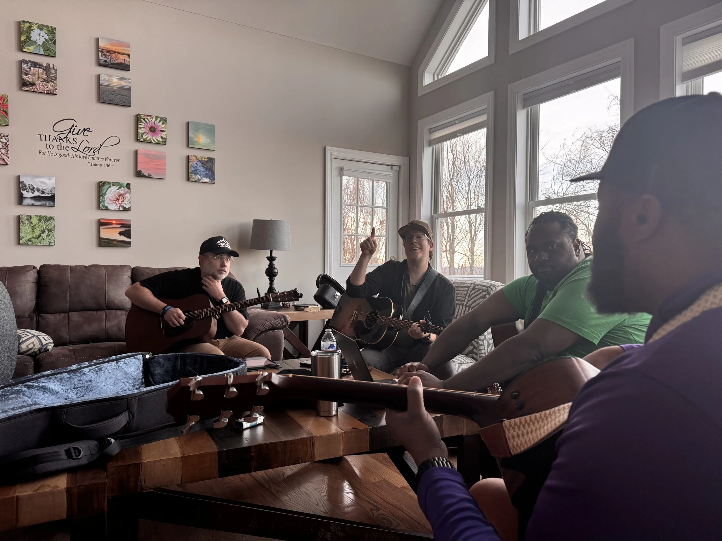 Four men in a living room, three playing guitars and one listening, with large windows and wall art.