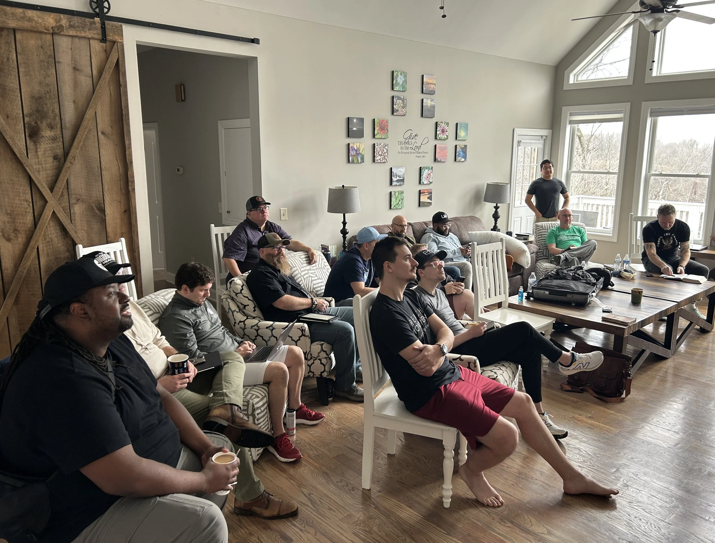 A group of people gathered in a living room, sitting on various chairs and couches, attentively watching a presentation or speaker. The room is bright with large windows and has hardwood floors with casual furniture and wall decorations.