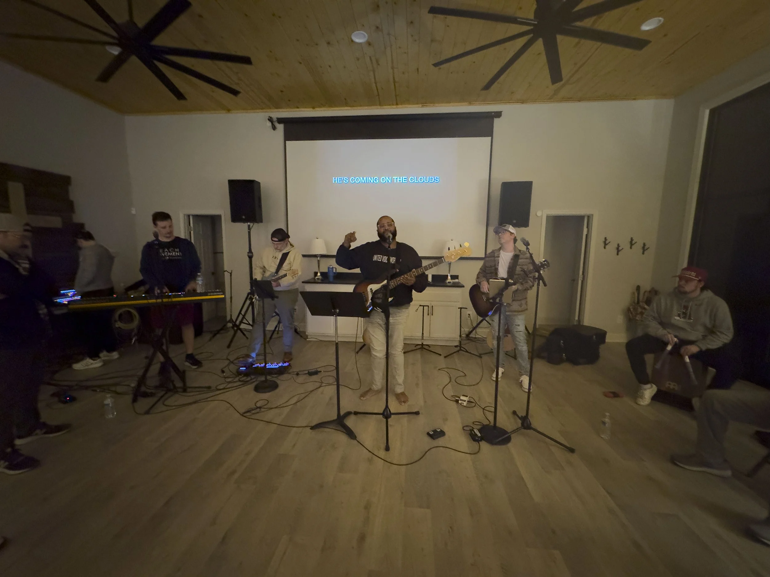 A group of seven people performing music in a cozy indoor space with a wooden ceiling and large windows. Two musicians in the center are playing guitars, and the man in the middle is singing with a microphone. Others are on instruments or relaxing, w