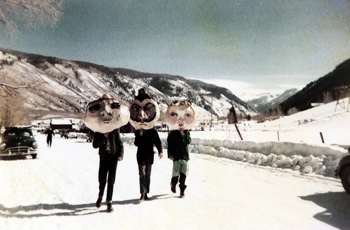 Three people wearing oversized papier-mâché heads walk along a snowy mountain road during Aspen’s Wintersköl celebration, with vintage cars and snow-covered hills in the background.