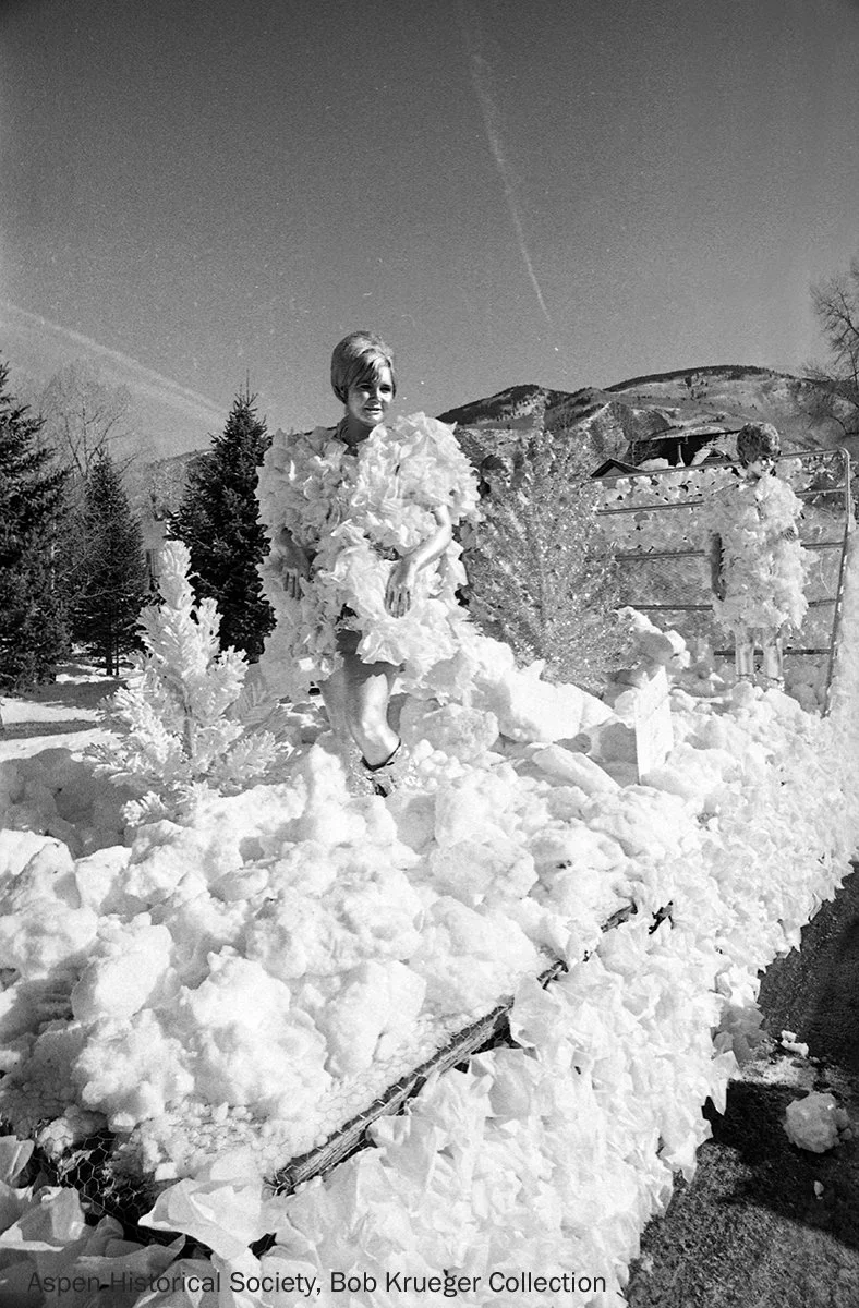  A vintage photo of a winter parade float decorated with artificial snow carrying two children dressed in fluffy white costumes, set against Aspen’s snowy landscape and mountains. 