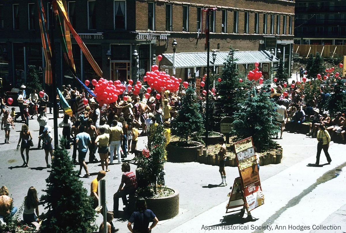  A lively crowd gathers in downtown Aspen with clusters of red balloons, celebrating in a pedestrian mall surrounded by storefronts and greenery 