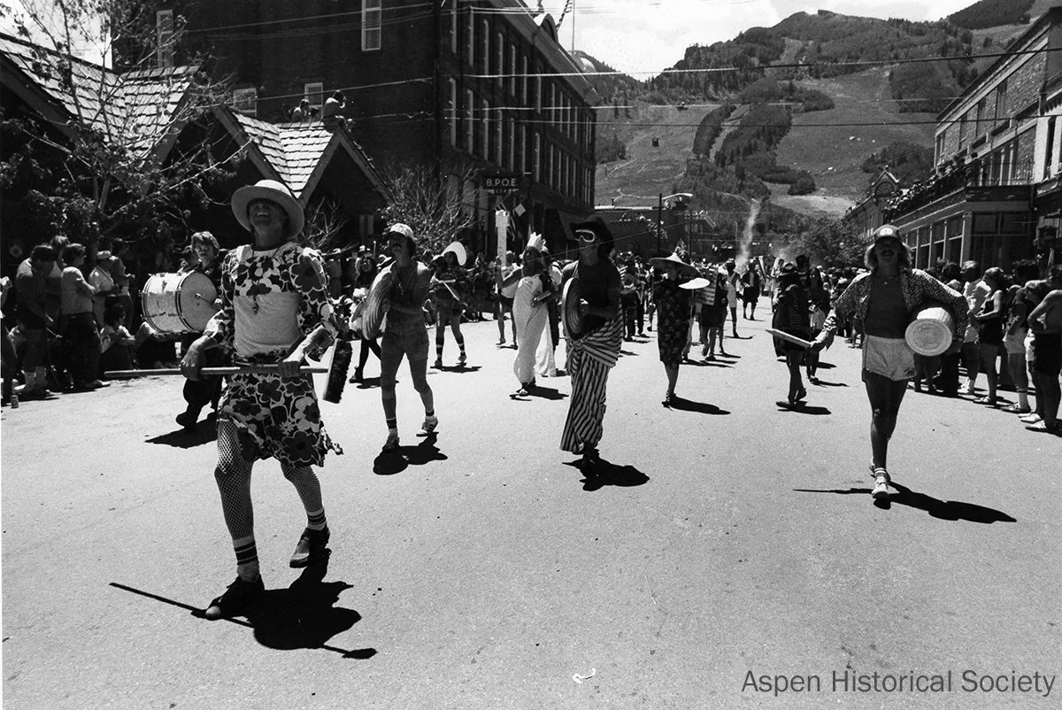  Vintage photograph of parade performers in playful, mismatched costumes marching down a sunny Aspen street, playing instruments as spectators line the sidewalks with mountain slopes in the background. 