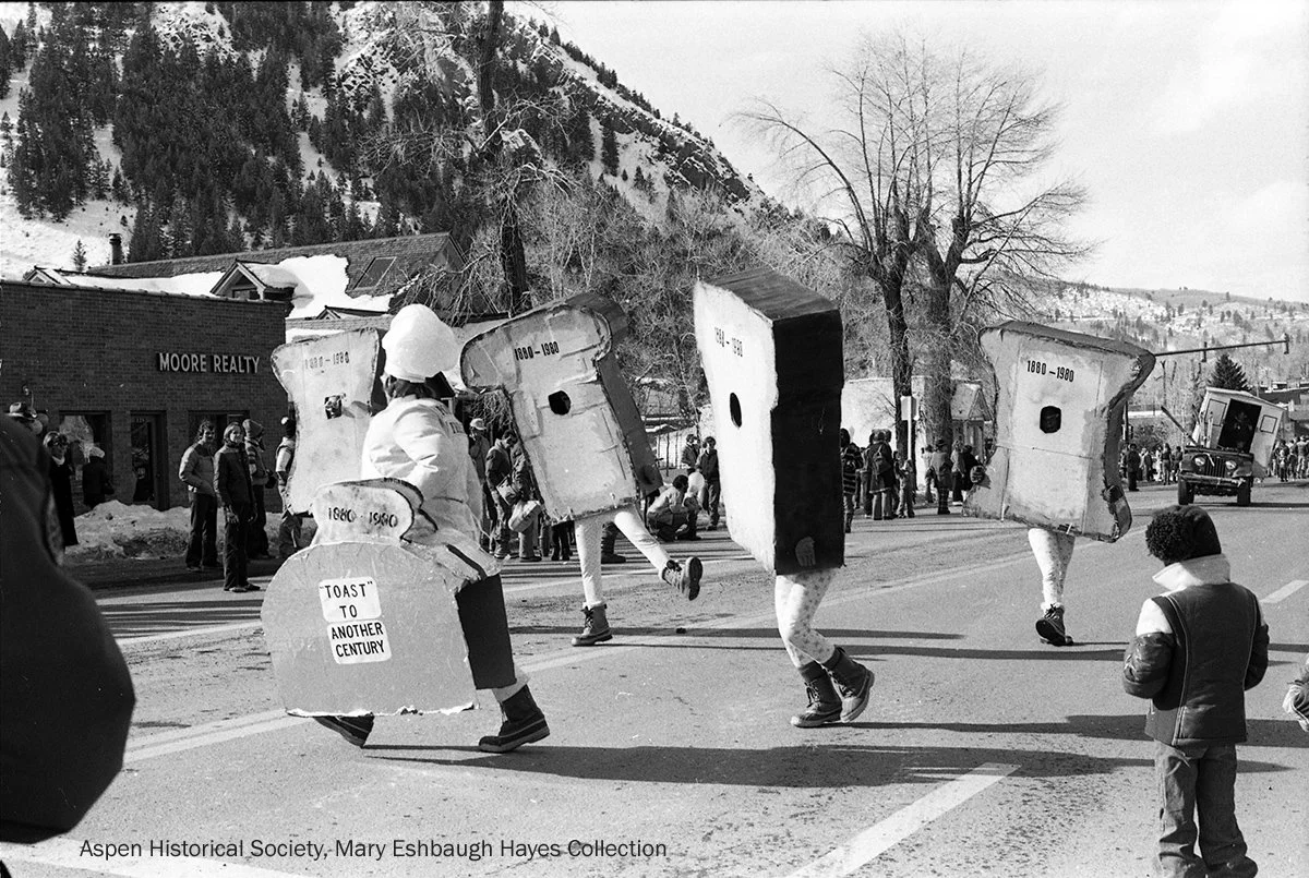  Vintage photograph of parade participants dressed as oversized slices of bread marching down a street during Aspen’s Wintersköl celebration, with onlookers gathered along the roadside. 