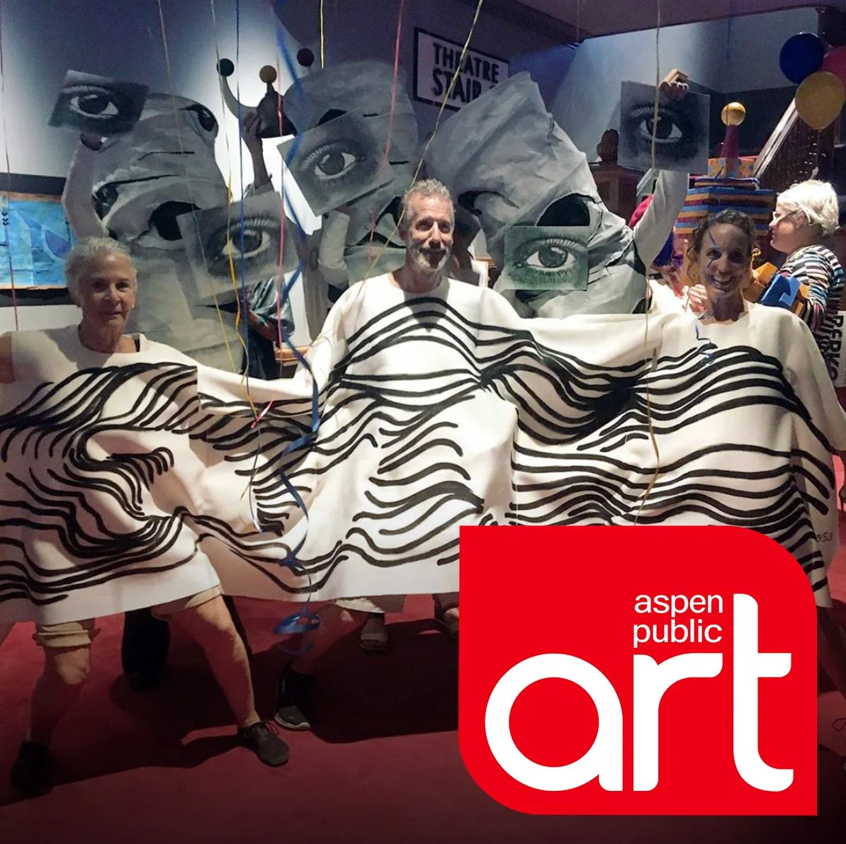 Aspen Public Art logo imposed atop a photo of three artists wearing a creative white tunic decorated with Bauhaus-inspired wavy black lines as an example of a creative art-themed costume