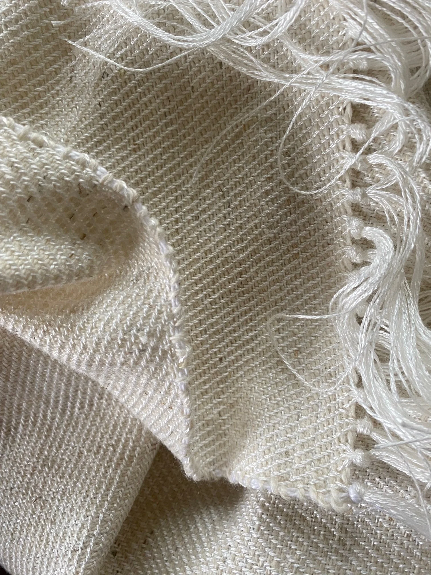 Hand-woven cream textile with knotted fringe and loose threads, revealing the structure and finishing of a woven piece.