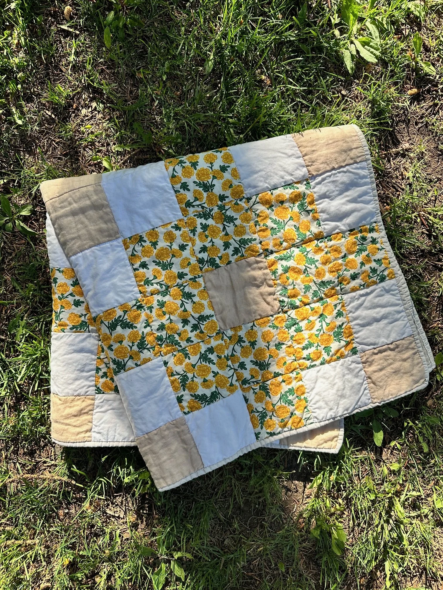 Small patchwork quilt with yellow floral fabric and neutral cotton blocks arranged in a geometric pattern, photographed outdoors on grass.