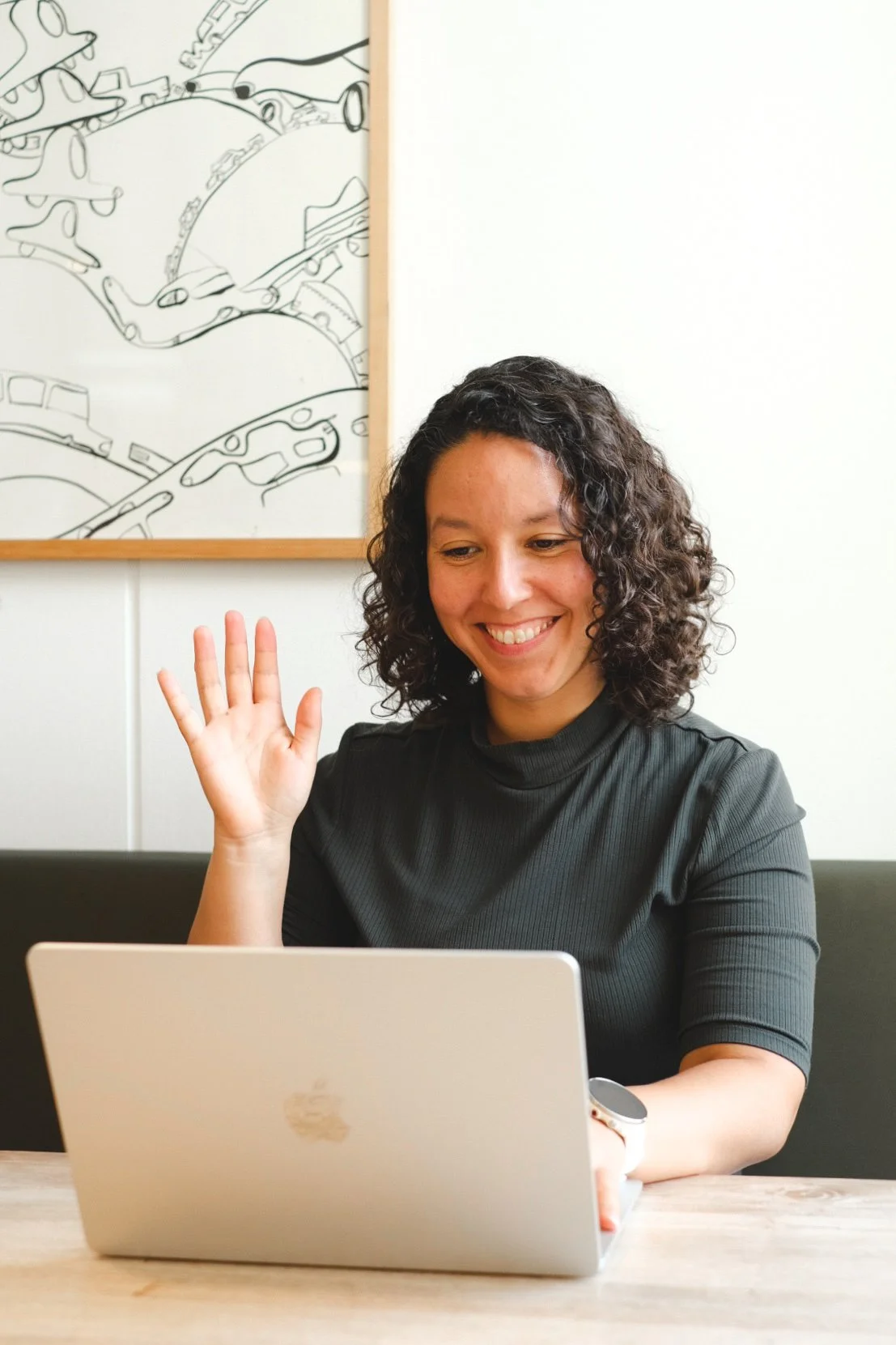 A woman with curly hair, wearing a black shirt, sitting at a table with a silver laptop, smiling and waving at the screen.