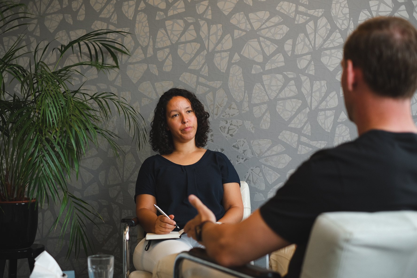 A woman sitting in a chair, holding a pen and a notepad, during a conversation with a man in a therapy or counseling session in a room with a decorative patterned wall and a potted plant.