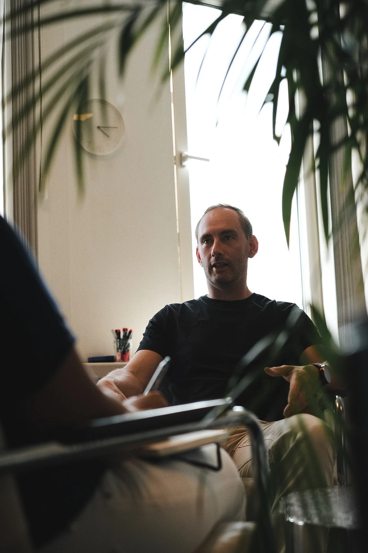 Man in black shirt sitting in an office or meeting room, engaged in conversation with another person, with sunlight coming through an open window behind him, surrounded by indoor plants.