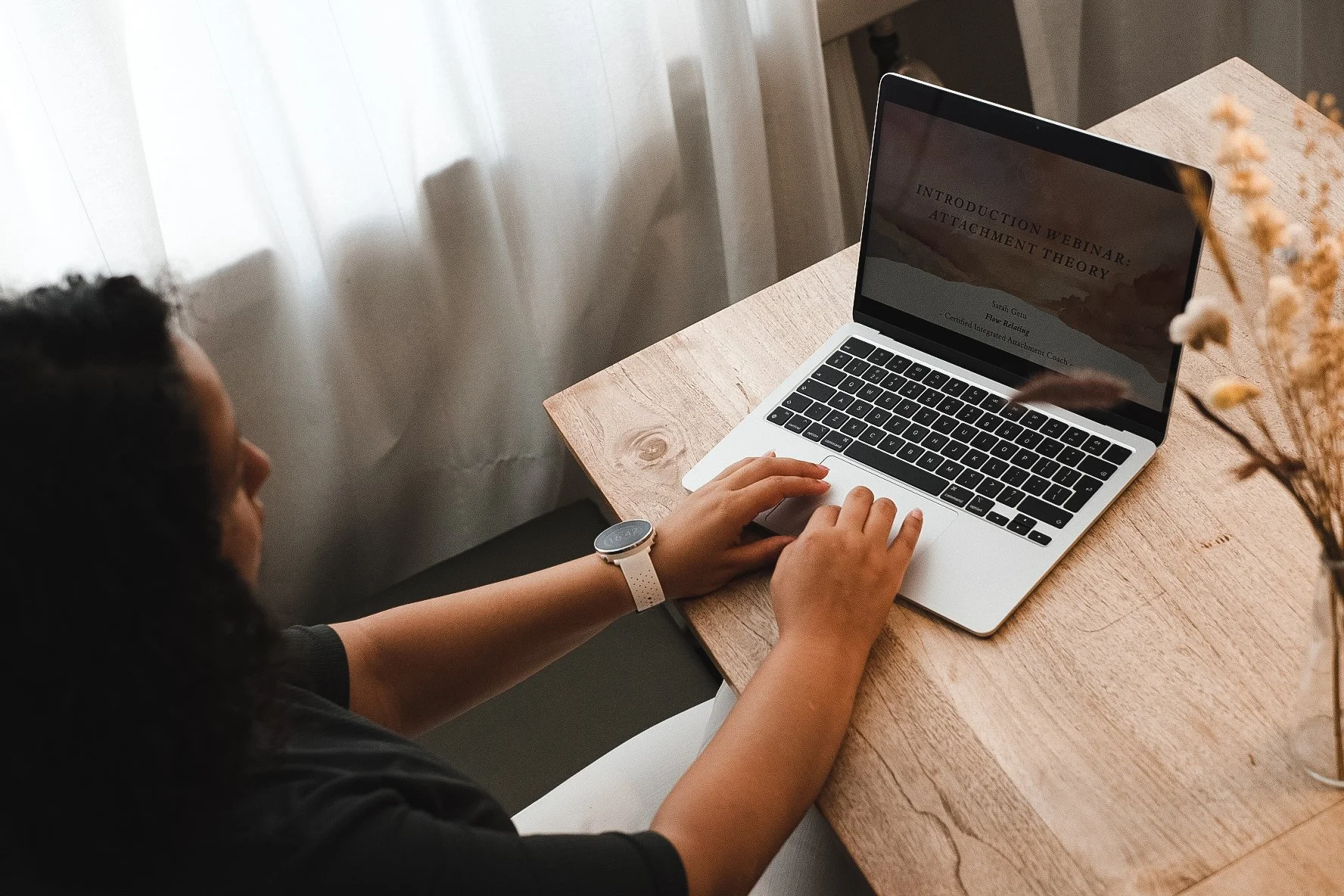 A person with dark, curly hair and wearing a white watch is sitting at a wooden table and looking at a silver laptop with a black keyboard. The laptop screen displays text for a webinar titled 'Introduction Webinar: Attachment Theory.' There are light-colored curtains in the background and a blurred vase of dried flowers on the right side of the table.