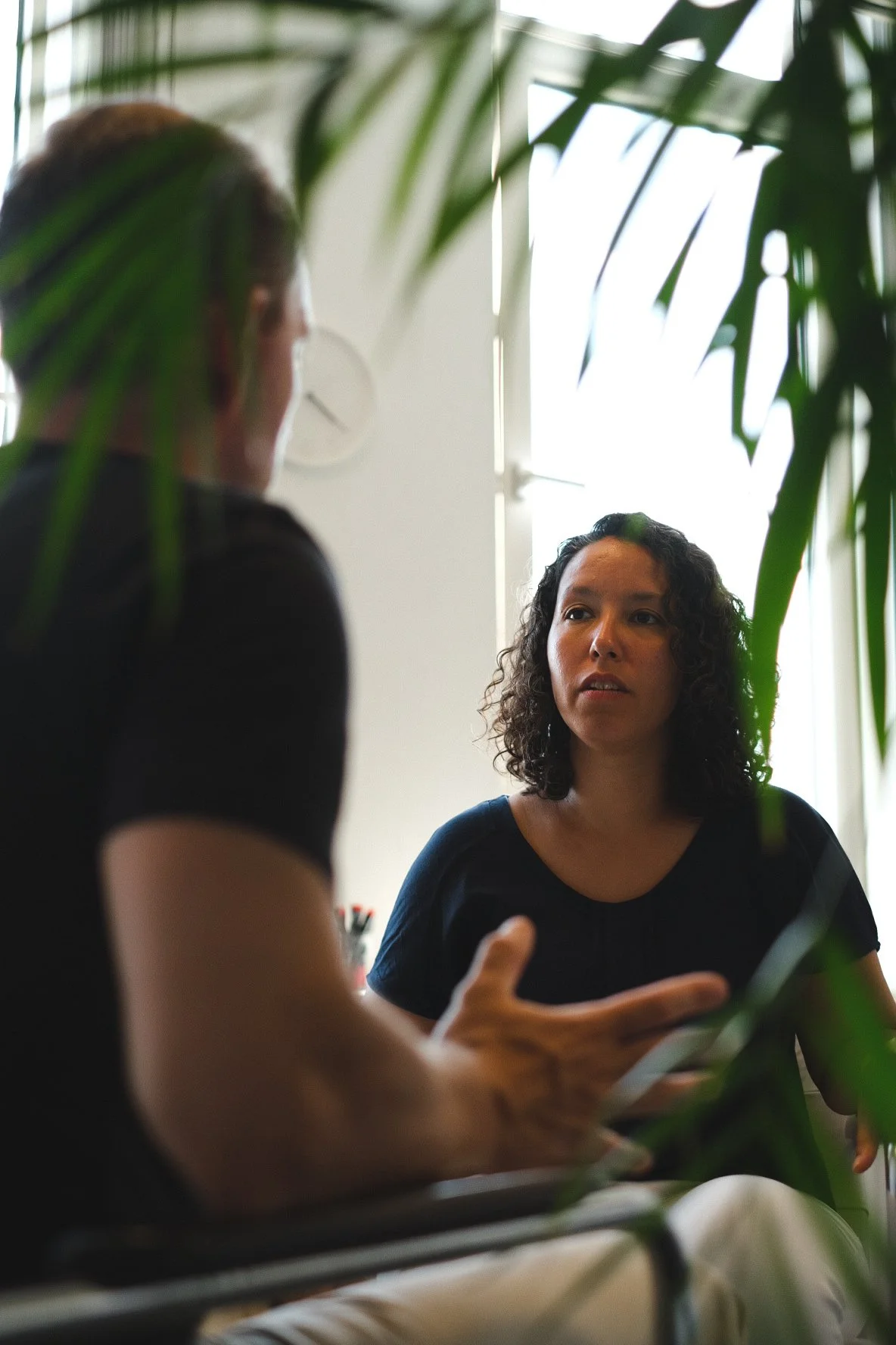 Two women engaged in a conversation in an office, with plants partially obscuring the view and sunlight coming through a window.
