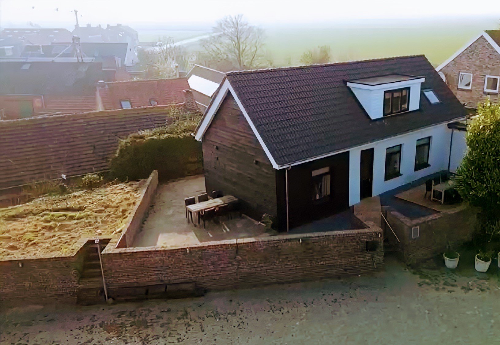Luchtfoto van een huis met donkere zijmuur en witte voorkant, omringd door een tuin met een terras en tafel, naast andere gebouwen.