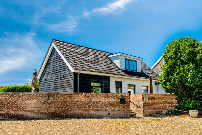 Een modern huis met een dak van grijze tegels, omgeven door een bakstenen muur en een grote groene boom, onder een blauwe hemel.