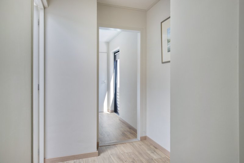 Binnenkijkend in een wit geschilderde gang met vloeren van licht hout, een deur en een raam bij de gang die licht binnen laat.