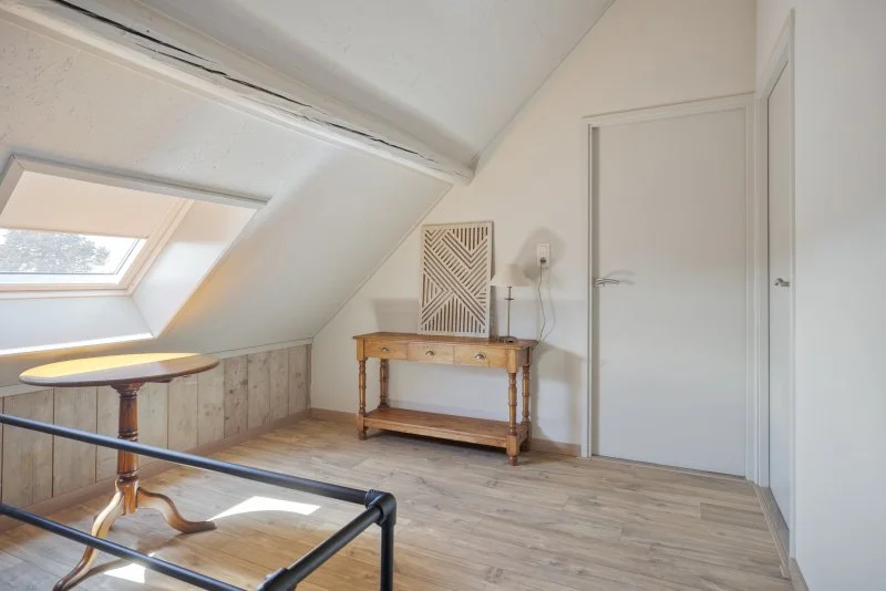 Een minimalistisch ingerichte zolderkamer met een houten tafel, een houten kast, een raam in het dak, en een witte deur.