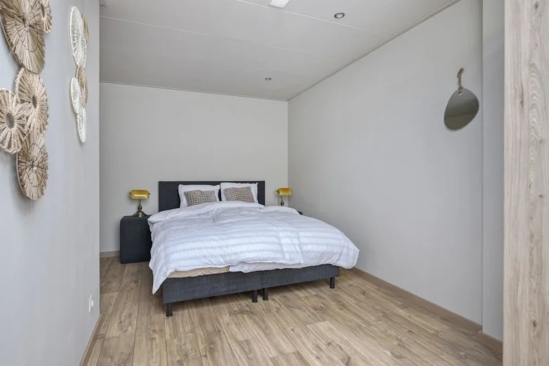 Een slaapkamer met een bed, twee nachttafels met lampen, witte beddengoed en decoratieve kussens, witte muren en een houten vloer.