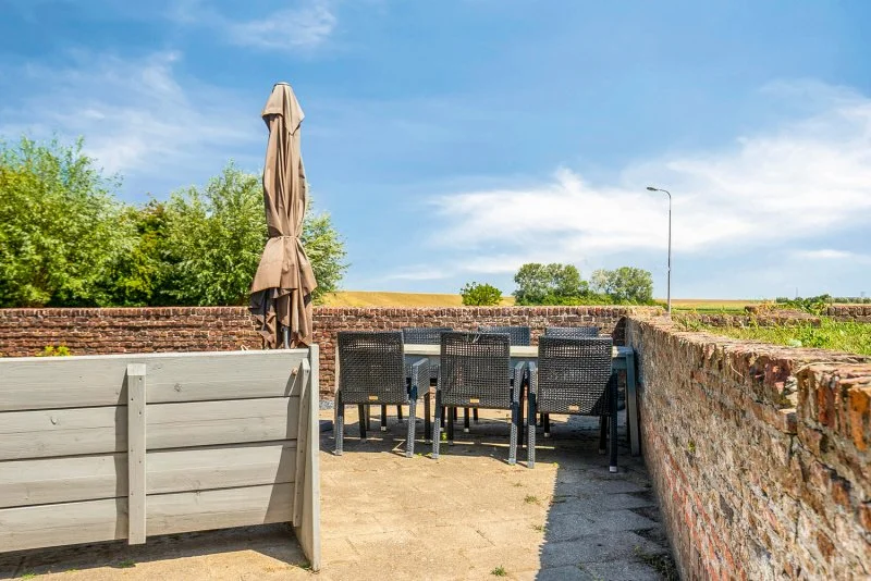 Buiten eetplek met tafel, toelen en een gesloten parasol op het terras van het 2 persoon vakantiehuis te huur bij Kloosterzande omringd door een lage muur, met groene bomen en een heldere blauwe hemel op de achtergrond.