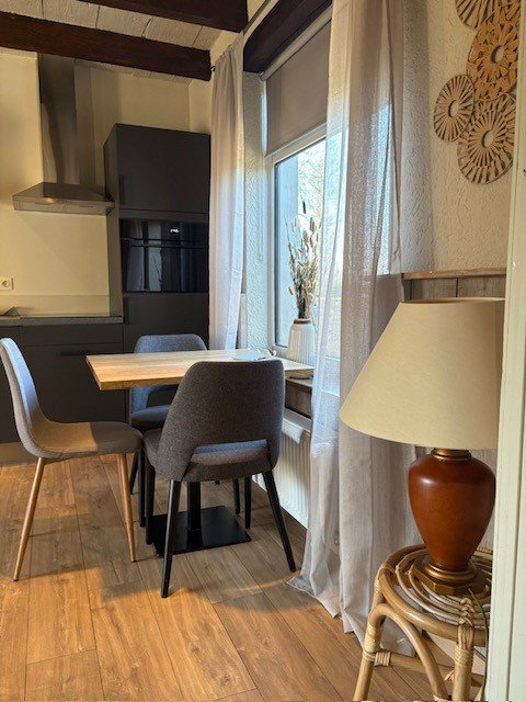 Eetkamer met houten tafel, vier grijze stoelen, lamp op rotan tafel, grote ramen, en keuken met donkere kasten en afzuigkap.