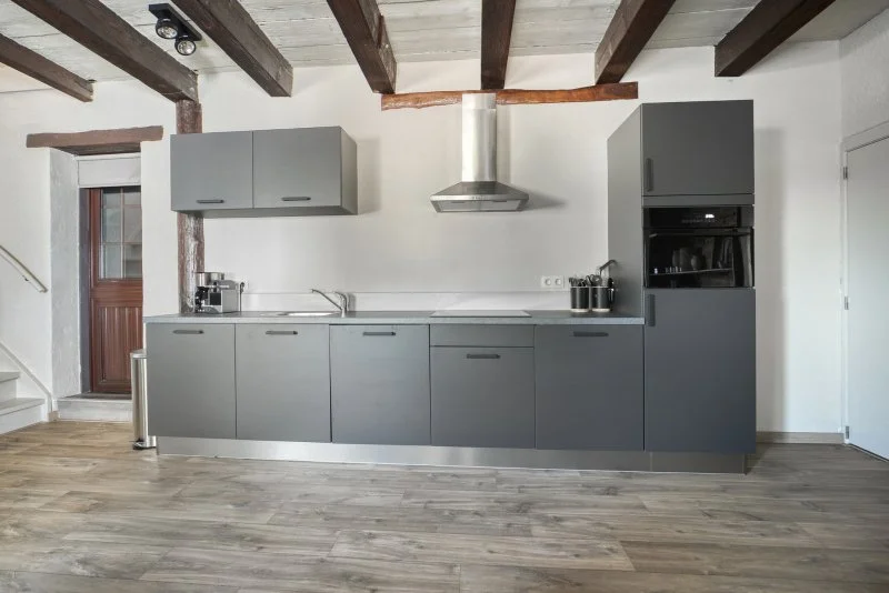 Een moderne keuken met grijze kasten, een ingebouwde oven, afzuigkap, en houten plafondbalken in een woonkamer.