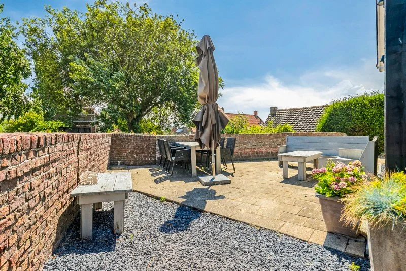 Het terras van het 2 personen vakantiehuisje in de buurt van Kloosterzande met een bakstenen muur, een tafel met vier stoelen en een gesloten parasol. Daarnaast een zitbank en een grote plantenbak met bloemen.
