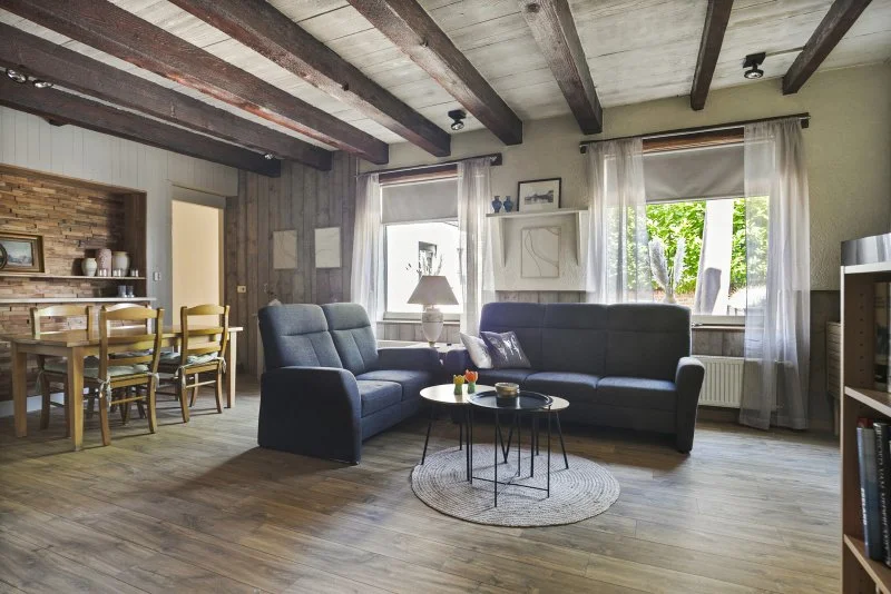 Gezellig woonkamer met twee blauwe banken, ronde salontafel, grote ramen met witte gordijnen, houten plafond en een eetkamer met houten tafel en stoelen op de achtergrond van het 6 persoon vakantiehuis te huur bij Paal Zeeuws Vlaanderen.