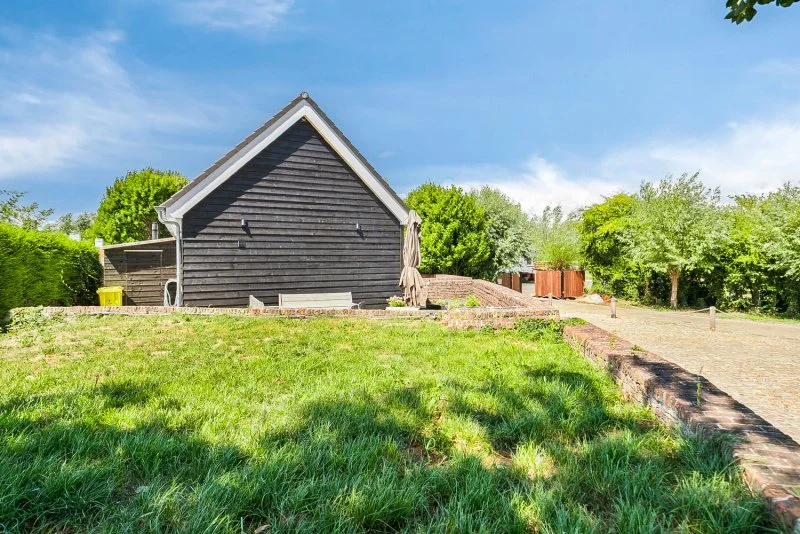 Zwart houten huis met een groene tuin en bomen, een oprit en een blauwe hemel.