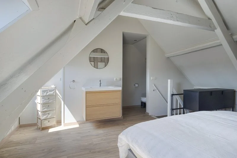 Slaapkamer met wit plafond, houten vloer en een bed met wit beddengoed. Aan de muur hangt een spiegel en er is een badkamermeubel van het 2 persoon vakantiehuis te huur bij Paal, Kloosterzande, Hulst.