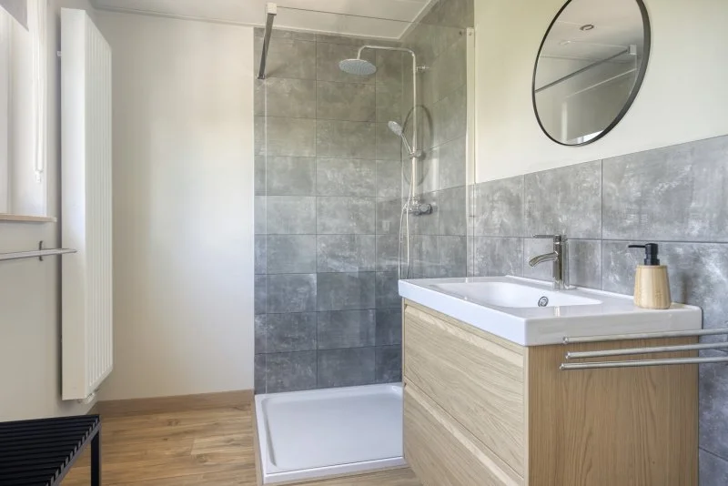 Moderne badkamer met inloopdouche, ronde spiegel, wastafel met houten onderkast en muurschilderingen, houten vloer en radiator.