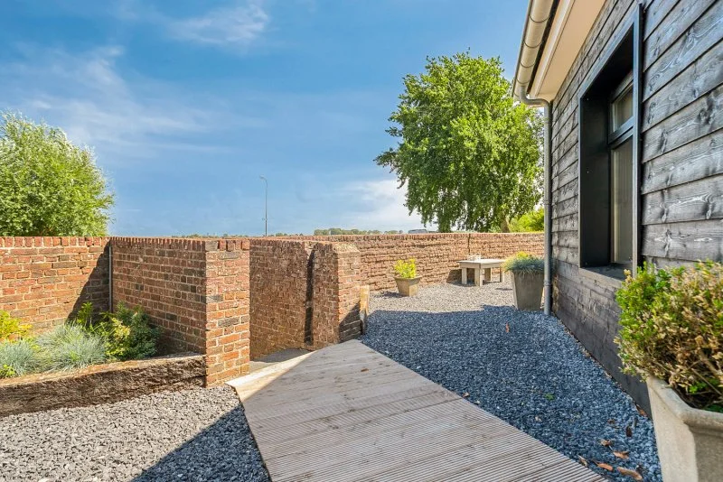 Buitenruimte met houten terras, grindpaden, plantenbakken en een bakstenen muur naast een houten huis onder een blauwe lucht.