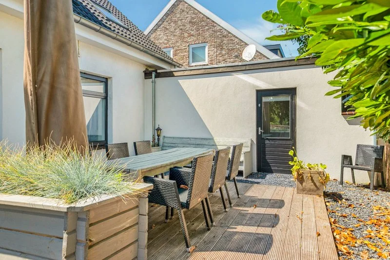 Buitenterras met een lange tafel en acht rieten stoelen onder een blauwe lucht, omgeven door planten en een muur van het vakantiehuis Den Burtman bij Paal