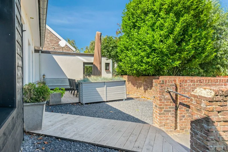 Een tuin met tuinmeubilair, plantenbakken, een grote boom en een betegeld pad onder een blauwe hemel.